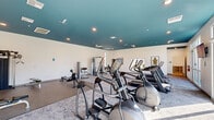 This modern fitness center is thoughtfully designed across three interconnected areas, offering a comprehensive workout environment. The main exercise floor is a generously sized, open space distinguished by a bold teal ceiling and recessed lighting that creates an energizing atmosphere. Large windows along the perimeter flood the room with natural light while providing views of the surrounding exterior. The main floor is outfitted with an extensive array of cardio equipment, including multiple treadmills, elliptical trainers, and a rowing machine, all arranged on patterned carpet. A dedicated strength training zone features rubberized flooring, a multi-station cable machine, a weight bench, and additional resistance equipment. A separate cycling and activity studio offers light wood-look flooring, spin bikes, stability balls, resistance bands, and open wall space, with floor-to-ceiling glass doors that connect to an outdoor area. An adjoining flex room with large windows and soft carpeting serves as a children's play area or quiet retreat, furnished with a low storage unit and colorful wall art. Throughout the facility, clean white walls, modern finishes, and ample ventilation contribute to a bright, functional, and welcoming fitness environment.
