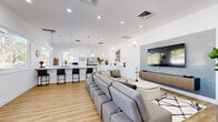 This thoughtfully designed single-floor space offers a seamless open-concept layout that combines functional work and social environments with a clean, contemporary aesthetic. Light hardwood flooring flows throughout, complementing the crisp white walls and recessed lighting that keep the space feeling bright and airy.

The central lounge area serves as the heart of the space, anchored by a large sectional sofa and modular seating arranged around a bold accent wall finished in textured concrete-look material, upon which a wall-mounted flat-screen TV and sleek floating media console are installed. A geometric patterned area rug and lush indoor plants add warmth and character.

Adjacent to the lounge, the kitchen area is a standout feature, boasting an expansive white quartz island with bar seating, white shaker cabinetry, stainless steel appliances, and pendant lighting. A dedicated galley-style prep kitchen runs alongside, offering additional counter space, cabinetry, a sink, and a large window with outdoor views.

Toward the rear of the space, frosted glass-paneled private offices or meeting rooms provide a sense of separation while maintaining visual openness. Natural light pours in through multiple windows, offering views of surrounding greenery. The overall ambiance is polished, professional, and welcoming.