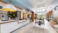Located at 1350 Gage St. in Columbus, Ohio, this thoughtfully designed single-floor community lounge offers a generous, open layout that seamlessly blends multiple functional zones into one cohesive space. The interior is defined by soaring double-height ceilings in the main reception area, floor-to-ceiling arched windows that flood the space with natural light, and an inviting palette of soft blues, warm wood tones, and rich teal cabinetry throughout.

A tiled reception counter with white subway tile detailing anchors the entry, while a dedicated coffee bar — framed by a decorative arched alcove, hexagonal tile flooring, and teal cabinetry with brass hardware — provides a welcoming refreshment station. Distinct seating zones cater to a variety of uses: a relaxed lounge area features tufted velvet chairs and a leather banquette beneath a wood-paneled ceiling, overlooking outdoor pool views through a large picture window; a more social area offers marble-topped round tables with modern chairs; and a bar-height counter with stools lines a sun-drenched window wall, ideal for individual work or casual conversation.

Decorative details such as wall-mounted art, open teal shelving, textured column accents, and curated lighting fixtures — including oversized pendant lights with gold interiors — contribute to the space's vibrant yet refined character. The combination of light wood plank flooring and hexagonal tile throughout ties the varied zones together with visual continuity.