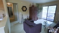 This single-floor, two-bedroom, one-bathroom residence offers a practical and comfortable layout designed for everyday living. The living room serves as the heart of the home, featuring plush wall-to-wall carpeting that adds warmth underfoot. Neutral-toned walls provide a versatile backdrop, complemented by a large sliding glass door that floods the space with natural light and opens to an outdoor balcony, offering views of lush greenery beyond. The living area provides ample room for seating and storage furniture, as demonstrated by the generous armchair and a substantial wood armoire that anchors the space. Decorative touches, including an octagonal mirror and ornamental accents, lend character to the walls. A hallway visible from the living room connects to the remainder of the unit, including the two bedrooms and bathroom, ensuring a straightforward and functional flow throughout. The overall ambiance is cozy and inviting, with natural light and greenery—both indoors and out—contributing to a relaxed, homey atmosphere well-suited for comfortable daily living.