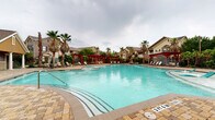 This outdoor amenity space centers around a generously sized freeform swimming pool with vivid turquoise water, designed to evoke a resort-like atmosphere. The pool features a gradual entry with built-in in-water lounge chairs, ideal for relaxing while partially submerged, as well as clearly marked depth indicators along the tiled pool edge. The surrounding deck is finished with an attractive paver pattern, providing ample space for lounging and socializing.

Several bold red pergolas are positioned around the perimeter, offering shaded seating areas furnished with lounge chairs, tables, and chairs — perfect for outdoor gatherings. Tall palm trees are interspersed throughout the landscape, enhancing the tropical ambiance of the space. Lush greenery and mature trees line the borders, providing a sense of privacy and enclosure.

A stone-clad pool house anchors one end of the space, featuring a covered patio with additional seating and serving as a practical hub for pool amenities. The surrounding multi-story residential buildings, finished in neutral earth tones with pitched roofs, are visible in the background, framing the outdoor area. The layout of the entire pool deck is thoughtfully designed to maximize both open recreational space and comfortable, shaded retreat areas.
