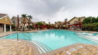 This outdoor amenity space centers around a generously sized freeform swimming pool with vivid turquoise water, designed to evoke a resort-like atmosphere. The pool features a gradual entry with built-in in-water lounge chairs, ideal for relaxing while partially submerged, as well as clearly marked depth indicators along the tiled pool edge. The surrounding deck is finished with an attractive paver pattern, providing ample space for lounging and socializing.

Several bold red pergolas are positioned around the perimeter, offering shaded seating areas furnished with lounge chairs, tables, and chairs — perfect for outdoor gatherings. Tall palm trees are interspersed throughout the landscape, enhancing the tropical ambiance of the space. Lush greenery and mature trees line the borders, providing a sense of privacy and enclosure.

A stone-clad pool house anchors one end of the space, featuring a covered patio with additional seating and serving as a practical hub for pool amenities. The surrounding multi-story residential buildings, finished in neutral earth tones with pitched roofs, are visible in the background, framing the outdoor area. The layout of the entire pool deck is thoughtfully designed to maximize both open recreational space and comfortable, shaded retreat areas.