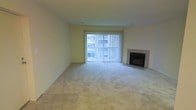 This single-floor space presents a clean, functional layout centered around a spacious living area. The room is finished with wall-to-wall carpeting in a neutral beige tone, complemented by tiled flooring near the entryway, creating a subtle transition between zones. Smooth, light-colored walls give the space a bright and airy feel throughout.

A standout feature of the living area is the tile-surround fireplace positioned along one wall, providing both a functional heating element and a classic focal point for the room. Adjacent to the fireplace, a large sliding glass door spans a significant portion of the wall and is dressed with vertical blinds that allow for adjustable natural light control. The door brings in ample daylight, brightening the interior and offering a view of the surrounding exterior.

The overall ambiance is understated and neutral, with a blank-canvas quality that lends itself well to a variety of furnishing and décor styles. The absence of clutter and the open floor plan allow the architectural elements — the fireplace and the generous glazed opening — to take center stage, making this a versatile and inviting living space.