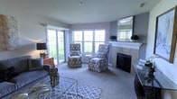 This single-floor residence offers a well-appointed layout designed for comfortable living. The living room serves as the centerpiece of the home, featuring a tile-surround fireplace with a white mantel, soft neutral-toned carpet, and an abundance of natural light streaming through multiple double-hung windows that overlook lush outdoor greenery. A decorative mirror above the mantel and a patterned area rug anchored by a glass coffee table contribute to the room's polished aesthetic. Two boldly patterned high-back accent chairs complement a plush gray sofa, creating an inviting seating arrangement. Large-scale wall art adds a finishing touch to the space.

Adjacent to the living area, the dining space accommodates a round glass table with contemporary dark chairs beneath a modern pendant light fixture, with views into the kitchen providing an open, connected feel. The kitchen is efficiently designed with dark cabinetry, granite countertops, tile flooring, and stainless steel appliances including a French door refrigerator and range. A pass-through window connects the kitchen to the entry area, enhancing the home's overall flow and functionality.