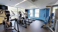 This fitness center offers a practical and inviting workout environment designed to support a range of cardio-focused exercise routines. The space is anchored by a set of treadmills and elliptical machines, providing residents or members with accessible options for daily fitness. A floor-to-ceiling glass window wall along one side of the room floods the space with generous natural light and offers views of mature trees outdoors, creating a refreshing backdrop during workouts. The interior features bold blue accent walls complemented by additional natural light from double-hung windows on the adjacent wall, keeping the atmosphere open and bright. Durable rubber flooring covers the workout area, providing both safety and comfort underfoot. A wall-mounted television is positioned for easy viewing from the cardio equipment. A seated exercise machine is tucked along the far wall, offering a low-impact strength or rehabilitation option. Overhead fluorescent lighting supplements the natural light, ensuring the space remains well-lit throughout the day. The overall layout is efficiently organized, maximizing the available floor space while maintaining an uncluttered, functional environment suited for daily exercise.