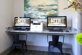 This single-floor business center offers a thoughtfully designed workspace that balances functionality with refined aesthetics. The room is anchored by a built-in granite countertop workstation accommodating two computer terminals and a multifunction printer, making it ideal for focused work sessions. Above the workstation, a large abstract painting in soft blues, greens, and yellows serves as a striking focal point, adding an artistic touch to the professional setting. The space features warm hardwood-style flooring throughout, complemented by neutral walls that create a calm, distraction-free atmosphere. A generously sized window with classic grid detailing floods the room with natural light and offers pleasant views of lush greenery and surrounding landscaping. The business center is accessed through a glass-paned French door, providing a sense of openness while maintaining a degree of privacy. A dedicated entry area with a granite ledge shelf is well-suited for displaying informational materials and brochures, enhancing the welcoming character of the space. An exterior glass door with a brick-paved corridor visible beyond reinforces the space's accessibility and its connection to the broader building environment. Overall, this business center combines practical amenities with elegant design details for a productive and comfortable experience.