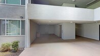 This ground-floor covered common area is part of a multi-story building, characterized by a clean and functional architectural design. The space features a large open layout beneath a substantial overhead structure, creating a sheltered transitional area that connects the building's interior to its exterior surroundings. The smooth concrete flooring extends throughout, providing a durable and easy-to-maintain surface. Ceiling-mounted flush lights offer even illumination across the space, ensuring visibility day or night. At the rear, a solid interior door provides direct building access, while an elevator is conveniently positioned to one side, facilitating easy vertical movement throughout the building. The stucco-finished walls and overhead soffit contribute to the structure's cohesive exterior aesthetic, presenting a neutral, light-toned palette throughout. A waste receptacle near the entrance reflects thoughtful amenity placement within the common area. The open-front design allows for natural airflow and light penetration, giving the space a bright and airy feel despite its sheltered nature. Overall, this area serves as a practical and well-appointed transitional zone, ideal for building entry, circulation, and everyday resident use.
