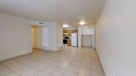 This single-floor space offers an open, functional layout where the living area flows directly into the kitchen. The floors throughout are covered in large, light-colored ceramic tiles that contribute to a clean and bright atmosphere. The walls are painted in a neutral off-white tone, keeping the interior feeling open and airy.

The kitchen is positioned toward the rear of the main living space and is equipped with warm wood-toned cabinetry, a white refrigerator, and a white range with an overhead ventilation hood. The cabinetry provides ample storage, and the layout allows for efficient use of the cooking area.

Adjacent to the kitchen, a doorway leads to an additional room that appears to include a closet or storage area with open shelving, making it well-suited for use as a bedroom or utility space. A separate doorway on the opposite side of the main room leads to another area, providing a degree of separation within the unit.

Ventilation is provided via ceiling and floor-level vents distributed throughout the space. Overhead lighting fixtures illuminate the interior uniformly. The overall design is practical and straightforward, prioritizing functionality and ease of maintenance.