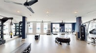 Located at 3010 Furr Court in Charlotte, NC, this well-appointed fitness center offers a generously sized, single-floor exercise space designed to support a wide range of workout routines. The interior features a clean, modern aesthetic with bold navy blue accent walls contrasting against crisp white ceilings and smooth, light-toned flooring that reflects the bright, airy atmosphere throughout.

The space is flooded with natural light through a series of large windows along the perimeter walls, each adorned with branded frosted decal accents. Multiple ceiling fans are distributed throughout, ensuring comfortable airflow during intense workouts. Recessed lighting supplements the natural light for consistent illumination across the entire floor.

Equipment is thoughtfully arranged to maximize usability and circulation. The cardio zone includes treadmills, stationary bikes, and a rowing machine, while the strength training area is equipped with multi-station cable machines, weight benches, and free weight racks stocked with kettlebells and dumbbells. Functional training is well-represented with TRX suspension systems, a heavy training tire, medicine balls, and open floor space for dynamic movement. Structural columns with navy-painted bases are integrated throughout, adding architectural character while defining activity zones within the open layout.