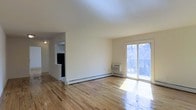 This single-floor space offers a clean, open layout characterized by light-toned hardwood floors that reflect natural light throughout the interior. The living area is spacious and uncluttered, with neutral cream-colored walls that contribute to a bright and airy atmosphere. A sliding glass door dominates one wall, providing direct access to an outdoor balcony and framing views of mature surrounding trees. The door serves as the primary source of natural light, flooding the living space with daylight.

Heating is provided by baseboard units running along the perimeter walls, and a wall-mounted air conditioning unit positioned near the sliding door ensures year-round climate comfort. The open-concept design allows for a clear sightline from the living area into an adjacent kitchen space, which is partially visible through a pass-through opening. The kitchen features dark appliances, including what appears to be a refrigerator, and is separated by a partial wall, maintaining a sense of distinction between the two areas while preserving the overall open feel of the layout. The space presents a functional and well-lit environment suitable for everyday living.