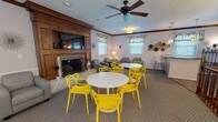 Located at 865 Revere Village Ct in Dayton, Ohio, this single-floor community space is thoughtfully designed to accommodate a variety of social gatherings and events. The main lounge area serves as the centerpiece, anchored by a classic wood-paneled fireplace fitted with a wall-mounted flat-screen TV above the mantle. A ceiling fan overhead ensures comfortable airflow throughout the carpeted room. The lounge is furnished with plush gray seating, including a sofa and armchairs, complemented by vibrant yellow accent chairs arranged around white round tables — creating a lively, welcoming atmosphere. Crown molding and wainscoting add a touch of architectural elegance throughout, while patterned valance window treatments allow natural light to filter in. A separate dining area provides additional seating with its own round table and yellow chairs, enhanced by wall sconces and views to adjoining rooms. The kitchenette is well-appointed with warm wood cabinetry, granite-style countertops, a sink, and small appliances — ideal for event preparation. The cohesive yellow, gray, and white color palette, along with decorative wall accents, gives the space a modern yet inviting character perfectly suited for community gatherings.