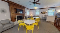 Located at 865 Revere Village Ct in Dayton, Ohio, this single-floor community space is thoughtfully designed to accommodate a variety of social gatherings and events. The main lounge area serves as the centerpiece, anchored by a classic wood-paneled fireplace fitted with a wall-mounted flat-screen TV above the mantle. A ceiling fan overhead ensures comfortable airflow throughout the carpeted room. The lounge is furnished with plush gray seating, including a sofa and armchairs, complemented by vibrant yellow accent chairs arranged around white round tables — creating a lively, welcoming atmosphere. Crown molding and wainscoting add a touch of architectural elegance throughout, while patterned valance window treatments allow natural light to filter in. A separate dining area provides additional seating with its own round table and yellow chairs, enhanced by wall sconces and views to adjoining rooms. The kitchenette is well-appointed with warm wood cabinetry, granite-style countertops, a sink, and small appliances — ideal for event preparation. The cohesive yellow, gray, and white color palette, along with decorative wall accents, gives the space a modern yet inviting character perfectly suited for community gatherings.