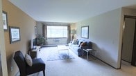 This single-floor space features a well-appointed living room that serves as its central gathering area. The room is finished with wall-to-wall carpeting in a neutral tone, contributing to a warm and understated atmosphere. A large window fitted with both a roller shade and floor-length curtains allows ample natural light to fill the space while offering flexible light control. Below the window, a baseboard heating unit runs along the wall, providing an efficient heating solution.

The living area is furnished with a grey upholstered sofa accented with decorative throw pillows, complemented by a glass-top coffee table resting on a patterned area rug. A side table with a slim metal frame adds a contemporary touch. A wall-mounted flat-screen television sits atop a wooden media console along the left wall, while a potted floor plant introduces a natural element to the room. Framed artwork on the walls adds visual interest without overwhelming the clean, neutral palette. The overall ambiance is calm and livable, with a layout that makes efficient use of the available space.