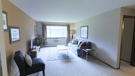 This single-floor space features a well-appointed living room that serves as its central gathering area. The room is finished with wall-to-wall carpeting in a neutral tone, contributing to a warm and understated atmosphere. A large window fitted with both a roller shade and floor-length curtains allows ample natural light to fill the space while offering flexible light control. Below the window, a baseboard heating unit runs along the wall, providing an efficient heating solution.

The living area is furnished with a grey upholstered sofa accented with decorative throw pillows, complemented by a glass-top coffee table resting on a patterned area rug. A side table with a slim metal frame adds a contemporary touch. A wall-mounted flat-screen television sits atop a wooden media console along the left wall, while a potted floor plant introduces a natural element to the room. Framed artwork on the walls adds visual interest without overwhelming the clean, neutral palette. The overall ambiance is calm and livable, with a layout that makes efficient use of the available space.