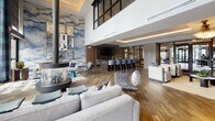Located at 360 Creamery Way in Exton, Pennsylvania, this upscale clubhouse lounge is a masterfully designed communal space that blends contemporary aesthetics with warm, inviting comfort. The open-plan layout flows seamlessly across hardwood-style flooring, uniting several distinct seating and gathering areas under soaring double-height ceilings accented with warm cove lighting.

At the heart of the space stands a striking cylindrical freestanding fireplace, flanked by sleek lounge seating and teal accent stools, all set against dramatic floor-to-ceiling grid-pane windows that flood the interior with natural light and frame views of the landscaped outdoor amenity area. A bold abstract mural in soft blue and white tones serves as a focal point for one of the lounge zones, complemented by a wall-mounted flat-screen television and a fully appointed bar with dark cabinetry, granite countertops, and high-back upholstered bar seating.

The reception and kitchen area features rich granite island surfaces, dark wood cabinetry, and glass-partition walls that maintain visual connectivity throughout. Additional seating vignettes include plush navy sofas, sculptural accent chairs, and statement pendant lighting, contributing to an atmosphere that is both refined and functional. The overall ambiance is modern, polished, and welcoming.
