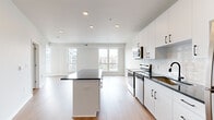 Located at 1636–1648 NW 38th St in Kansas City, MO, this thoughtfully designed single-floor residence combines contemporary finishes with a bright, open layout. The kitchen serves as a central highlight, showcasing crisp white cabinetry with matte black hardware, dark granite countertops, a center island with additional counter space, and a full suite of stainless steel appliances including a range, microwave, and refrigerator. A white subway tile backsplash adds a clean, modern touch. Warm hardwood-style flooring flows seamlessly throughout the entire unit, creating a cohesive and inviting atmosphere. The living area is generously proportioned, featuring a curved wall of large windows that flood the space with natural light and offer views of the surrounding neighborhood. The open entry area connects the kitchen and living spaces in a practical, free-flowing arrangement. The bathroom is finished to a high standard, featuring a walk-in glass-enclosed shower with large-format white tile, a dark granite vanity top, white cabinetry, and matching black hardware. A spacious walk-in closet provides substantial storage with built-in wire shelving and hanging rods. Recessed lighting throughout the unit ensures consistent illumination, complementing the clean, neutral color palette that defines the overall aesthetic of this well-appointed residence.