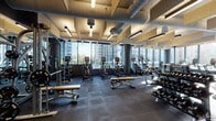 This contemporary fitness center is designed to deliver a premium workout experience within a sleek, urban setting. The space is defined by expansive floor-to-ceiling windows that wrap around the perimeter, flooding the interior with abundant natural light and offering striking views of the surrounding city skyline — creating an energizing backdrop for every workout.

The facility is organized into distinct training zones. A dedicated cardio area lines the window-facing walls, housing a robust selection of treadmills, ellipticals, stationary bikes, and stair climbers, all positioned to take full advantage of the outdoor views. A comprehensive strength training section features cable machines, multi-functional weight stations, adjustable benches, and a dumbbell rack, catering to a wide range of fitness routines.

Underfoot, durable rubber flooring in a dark tile pattern provides both safety and stability throughout the space. The ceiling showcases an industrial aesthetic with exposed ductwork, textured acoustic panels, and suspended linear lighting fixtures that complement the overall design language. Dark structural columns punctuate the floor plan, adding to the bold, modern atmosphere.

Entry to the facility is through a glass-paneled vestibule with dark metal framing, contributing to the polished, professional feel. Full-length mirrors along interior walls further enhance the sense of openness and allow users to monitor their form with ease.