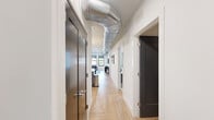 This thoughtfully designed single-floor residence offers two bedrooms and three bathrooms across a well-appointed layout. The open-concept kitchen and living area serves as the heart of the home, featuring two-tone cabinetry in grey and white, stainless steel appliances, a large island with a white countertop, and sculptural pendant lighting overhead. Light herringbone-pattern hardwood flooring flows throughout, adding warmth and visual continuity.

The living room is bright and inviting, benefiting from oversized windows that frame outdoor views and fill the space with natural light. A central hallway connects the main living area to the private quarters, with exposed metallic ductwork running overhead adding an industrial-modern design element. Dark-finished doors provide a striking contrast against the crisp white walls.

The primary bedroom is a comfortable retreat featuring a ceiling fan, a bold accent wall, and sliding glass doors opening to a private balcony with outdoor seating. A dedicated walk-in closet with built-in shelving and hanging rods provides generous storage. The bathrooms are finished with large-format marble-look tile, floating vanities, and glass-enclosed showers. A dedicated laundry room with side-by-side washer and dryer and overhead cabinetry completes the unit.