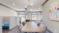 Located at 2550 W Ironwood Hill Dr in Tucson, Arizona, this single-floor cyber lounge and co-working amenity space offers a well-appointed environment designed for both focused work and casual collaboration. The space is defined by its elegant architectural details, including decorative crown molding, ornamental pilasters, and a series of tall arched windows that flood the interior with natural light and frame views of the surrounding Tucson landscape, including native desert vegetation and palm trees.

The flooring throughout features a stylish patterned carpet in blue-gray tones that anchors the contemporary furnishings. A central communal table with upholstered chairs serves as a collaborative hub, illuminated overhead by a striking mid-century-inspired brass and glass globe chandelier. Adjacent to this area, a dedicated computer workstation zone offers multiple desktop terminals arranged along a built-in desk, complemented by pendant lighting and leather-accented task chairs.

Private acoustic pods with built-in blue seating provide semi-enclosed spaces for focused individual work or small meetings. Adjustable wall-mounted task lamps add functional lighting throughout. The walls are painted in soft white and sage green tones, and curated artwork, including an abstract canvas, adds personality to the space. The overall ambiance is modern, welcoming, and well-suited for productivity.
