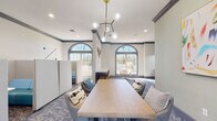 Located at 2550 W Ironwood Hill Dr in Tucson, Arizona, this single-floor cyber lounge and co-working amenity space offers a well-appointed environment designed for both focused work and casual collaboration. The space is defined by its elegant architectural details, including decorative crown molding, ornamental pilasters, and a series of tall arched windows that flood the interior with natural light and frame views of the surrounding Tucson landscape, including native desert vegetation and palm trees.

The flooring throughout features a stylish patterned carpet in blue-gray tones that anchors the contemporary furnishings. A central communal table with upholstered chairs serves as a collaborative hub, illuminated overhead by a striking mid-century-inspired brass and glass globe chandelier. Adjacent to this area, a dedicated computer workstation zone offers multiple desktop terminals arranged along a built-in desk, complemented by pendant lighting and leather-accented task chairs.

Private acoustic pods with built-in blue seating provide semi-enclosed spaces for focused individual work or small meetings. Adjustable wall-mounted task lamps add functional lighting throughout. The walls are painted in soft white and sage green tones, and curated artwork, including an abstract canvas, adds personality to the space. The overall ambiance is modern, welcoming, and well-suited for productivity.