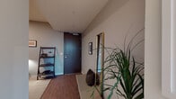 This thoughtfully designed single-floor residence showcases a contemporary aesthetic throughout, with clean lines, neutral tones, and high-quality finishes that create a cohesive and sophisticated atmosphere.

The entryway sets a welcoming tone, featuring rich wood-look flooring, a bold dark-paneled front door, a gold-framed full-length mirror, and a lean bookshelf that adds functional style. The space transitions smoothly into the main living area, where floor-to-ceiling windows frame impressive panoramic views of the surrounding cityscape and distant mountains, flooding the room with natural light. The living area features deep teal carpeting, curated wall art, and a comfortable seating arrangement ideal for relaxing or entertaining.

The kitchen is outfitted with flat-panel light wood cabinetry, stainless steel appliances, a gas range, and a bright white countertop with a subtle backlit under-cabinet design. A marble-topped dining table adds an elegant touch nearby.

The bedroom continues the floor-to-ceiling window theme, offering sweeping outdoor views alongside a wall-mounted television on a floating wood shelf. The bathroom is finished in warm natural stone tile, featuring a soaking tub, an illuminated vanity mirror, and light wood cabinetry.

A dedicated den or study provides an intimate, creative retreat with dark accent walls, open wooden shelving, curated artwork, and warm task lighting.