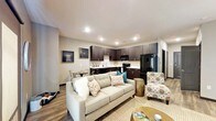 This single-floor residence offers a thoughtfully designed open-concept layout that seamlessly connects the living, dining, and kitchen areas into one cohesive and functional space. The living area is anchored by a plush, light-toned sofa accented with decorative throw pillows, complemented by a patterned accent chair and a warm area rug that adds texture to the rich hardwood-style flooring throughout. Recessed lighting in the smooth ceiling provides consistent, modern illumination across the entire space. The kitchen is a standout feature, showcasing dark espresso cabinetry that contrasts sharply with the light countertops and a clean white tile backsplash. Black appliances, including a full-size refrigerator, range, and microwave, lend the kitchen a sleek, contemporary feel. A compact dining area adjacent to the kitchen provides an intimate setting for meals. Neutral gray walls create a versatile backdrop that ties the design elements together, while wall art adds a personal touch. The overall ambiance is modern, warm, and inviting, with a well-balanced mix of dark and light tones that give the space a sophisticated yet comfortable character.