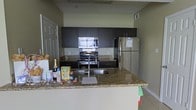 This single-floor space centers around a well-appointed kitchen that blends functionality with modern design. The kitchen features dark espresso-toned cabinetry that extends to the upper and lower sections, providing ample storage throughout. Granite countertops with warm, varied toning add a polished look and durable workspace. Stainless steel appliances, including a refrigerator, gas range, and over-the-range microwave, complement the contemporary aesthetic cohesively.

A peninsula with a built-in double sink extends outward, creating a natural divide between the kitchen and the adjacent living area. This peninsula also serves as a breakfast bar or casual dining surface, enhancing the space's versatility. Light tile flooring runs throughout, contributing to a clean, open feel. The walls are finished in a neutral green-gray tone, providing a subtle, calming backdrop that complements the dark cabinetry. Recessed overhead vents indicate a central HVAC system. Overall, the kitchen exudes a practical yet refined ambiance, well-suited for both everyday living and entertaining.