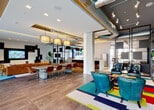 Located at 405 NE 2nd Street in the heart of Fort Lauderdale, this dynamic amenity floor delivers a bold, contemporary aesthetic that blends industrial design with vibrant, eclectic décor. The open-plan layout connects multiple distinct zones, each with its own character while maintaining a cohesive flow throughout the space. Exposed ductwork and track lighting run along the ceilings, complementing warm wood-look tile flooring and polished concrete surfaces to create an energetic, loft-inspired atmosphere. A central lounge area features teal leather armchairs arranged over colorful geometric rugs, anchored by open shelving units displaying curated décor. An adjacent media room offers a wall-mounted entertainment system framed by asymmetric display niches, with mid-century modern seating gathered around a central coffee table. A fully equipped kitchen and bar area includes stainless steel appliances, warm wood cabinetry, a spacious island with bar seating, and an herb planter detail. A large communal dining table beneath statement drum pendant lights seats a generous group, with floor-to-ceiling windows and glass doors opening to a poolside terrace beyond. A graphic feature wall adorned with bold, large-scale art and mounted screens adds a striking visual anchor, while a tucked-away lounge with built-in banquette seating offers a quieter retreat. A game area with a foosball table completes the entertainment offerings.