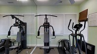 Located at 4545 Cary Glen Blvd in Cary, North Carolina, this fitness center offers a dedicated single-floor space designed to support a well-rounded workout routine. The room is appointed with light green walls that create a fresh, energizing atmosphere, complemented by natural light streaming in through multiple windows fitted with horizontal blinds. A full-length mirror spans one wall, visually expanding the space and providing a practical reference point for exercise form. The facility is equipped with a multi-function cable weight machine, ideal for a wide range of strength training exercises, as well as an elliptical trainer for low-impact cardiovascular workouts. Additional cardio and strength equipment is visible throughout the room, offering users a comprehensive set of training options. The flooring features durable, cushioned carpeting suited for gym use, and ceiling ventilation ensures comfortable air circulation during workouts. The overall ambiance is clean, functional, and focused, making this fitness center a practical and accessible amenity for residents or members seeking a convenient place to exercise.