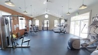 This fitness center is thoughtfully designed to offer a comprehensive workout environment in a bright, open setting. The space is defined by striking vaulted ceilings that rise to a central peak, creating an airy, expansive atmosphere enhanced by a combination of pendant lighting and ceiling fans for optimal comfort during exercise.

Natural light floods the room through an array of large windows along the walls, as well as a distinctive circular accent window near the peak of the vaulted ceiling, giving the space a welcoming and energizing feel. The durable dark rubber flooring provides both safety and practicality throughout.

The strength training area features a multi-function cable and weight machine, a free-weight rack with a full range of dumbbells, a barbell station with weight plates, and an adjustable workout bench — offering a complete setup for resistance training. On the cardio side, the space is outfitted with treadmills, an elliptical, and a stationary bike, along with wall-mounted screens for entertainment. A stability ball and additional accessories round out the amenities. The open floor plan allows for ample circulation and flexibility, making this fitness center a fully functional and inviting space for a wide range of exercise routines.