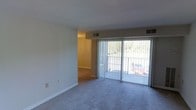 This single-floor residential space offers a clean, functional layout designed for comfortable everyday living. The main living area is a standout feature, highlighted by expansive sliding glass doors fitted with vertical blinds that flood the room with natural light while providing a view of the surrounding greenery outside. The space transitions smoothly between the carpeted living area and an adjoining room finished with hard flooring, suggesting a practical separation between living and dining or utility spaces. Neutral-toned walls and standard white trim throughout give the interior a bright, move-in-ready feel. Ceiling-mounted HVAC vents are evenly distributed, ensuring consistent climate control across the space. The overall ambiance is light and airy, supported by the generous window coverage and the openness of the layout. The connection between the interior and the outdoors enhances the sense of space, making the living area feel larger and well-ventilated. This straightforward, well-maintained space provides a solid foundation that is easy to personalize and adapt to a variety of living styles.