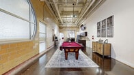 Located at 3031 Norfolk St in Richmond, VA, this single-floor commercial space offers a generously sized, open-plan layout that blends industrial character with modern functionality. The design retains its raw architectural roots through soaring exposed ceilings with visible structural trusses and mechanical systems, polished concrete floors, and original brick masonry walls — hallmarks of Richmond's adaptive reuse aesthetic.

The space is organized across a long, rectangular footprint that flows from a bright, open lounge area into a dedicated recreation zone. The lounge features a combination of polished concrete and terracotta tile flooring, high clerestory windows that allow abundant natural light, structural columns, and flexible seating arrangements suited for gathering or casual meetings. A bar-height counter with stools anchors one corner, adding a social hub to the area.

The recreation wing showcases a full-size billiards table as its centerpiece, surrounded by wall-mounted displays, a kitchenette with dark cabinetry, and decorative wall art. At the far end, an open event or flex area provides a clean, unobstructed canvas, lined with large windows and framed art pieces. A connecting corridor with additional framed artwork leads to private rooms and restrooms, giving the space a complete, well-rounded layout ideal for community, office, or event use.
