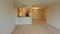 This single-story space offers a functional and open layout designed for comfortable everyday living. The living area is spacious and carpeted, with warm beige walls and clean white baseboards that give the space a neutral, welcoming feel. A textured ceiling adds subtle architectural character throughout.

A key feature of the main living area is a set of three evenly spaced windows along one wall, allowing generous natural light to fill the room while offering views of the surrounding exterior. A door with clean white trim is positioned to the side, providing convenient access to an adjacent space.

The kitchen is designed with a pass-through opening that connects it to the main living and dining areas, promoting an open flow between spaces. Wooden cabinetry, dark countertops, and black appliances furnish the kitchen, while recessed ceiling details add depth. A separate dining area is distinguished by an elegant chandelier, lending a touch of refinement to the otherwise casual setting.

Overall, the space presents a straightforward, practical design with consistent warm tones, plush carpeting throughout, and thoughtful layout choices that support both daily function and ease of movement.