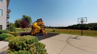 This outdoor recreational space offers a well-appointed play area designed to accommodate active use in a natural setting. The centerpiece is a substantial wooden playground structure equipped with a bright yellow tube slide, a ladder for climbing access, and a colorful canopy roof, all set atop a generous bed of dark rubber mulch that provides a safe, impact-absorbing surface around the equipment. Mature ornamental shrubs and trees, including a distinctive red-leafed variety, frame the perimeter of the play area, adding visual interest and a sense of enclosure without obstructing sightlines.

Directly adjacent to the playground, a smooth concrete surface serves as a dedicated basketball court, complete with painted court markings and a hoop, offering a versatile hard-surface activity zone that complements the softer play area nearby. The transition between the two zones is seamless, making the overall layout practical and easy to navigate.

The surrounding landscape opens up to expansive views of natural meadow grasses and a tree line in the distance, creating an open, park-like atmosphere under a clear blue sky. The combination of structured play equipment, sport court, and natural surroundings makes this space a comprehensive outdoor recreational area suited for a range of ages and activities.