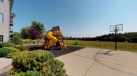 This outdoor recreational space offers a well-appointed play area designed to accommodate active use in a natural setting. The centerpiece is a substantial wooden playground structure equipped with a bright yellow tube slide, a ladder for climbing access, and a colorful canopy roof, all set atop a generous bed of dark rubber mulch that provides a safe, impact-absorbing surface around the equipment. Mature ornamental shrubs and trees, including a distinctive red-leafed variety, frame the perimeter of the play area, adding visual interest and a sense of enclosure without obstructing sightlines.

Directly adjacent to the playground, a smooth concrete surface serves as a dedicated basketball court, complete with painted court markings and a hoop, offering a versatile hard-surface activity zone that complements the softer play area nearby. The transition between the two zones is seamless, making the overall layout practical and easy to navigate.

The surrounding landscape opens up to expansive views of natural meadow grasses and a tree line in the distance, creating an open, park-like atmosphere under a clear blue sky. The combination of structured play equipment, sport court, and natural surroundings makes this space a comprehensive outdoor recreational area suited for a range of ages and activities.