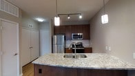 This single-floor space showcases a thoughtfully designed open-concept layout that centers around a well-appointed kitchen. The kitchen is a standout feature, highlighted by rich dark cabinetry that provides ample storage alongside a classic white subway tile backsplash. The expansive granite-topped island anchors the space and includes an undermount stainless steel sink with a modern arc faucet, offering both functionality and visual appeal. Pendant lighting fixtures hang gracefully above the island, complementing the track lighting overhead to create a warm, layered lighting scheme. The kitchen is fully equipped with a stainless steel refrigerator, a gas range, and a built-in microwave, making it both practical and contemporary in design. Light hardwood flooring runs throughout, adding warmth to the neutral color palette. The open layout allows the kitchen to flow naturally into the adjacent living areas, enhancing the sense of space and connectivity throughout the unit. A closet near the kitchen entry provides additional storage. Overall, the space presents a clean, modern aesthetic with quality finishes and functional design elements that make it both comfortable and inviting.