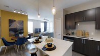 This single-floor residence showcases a thoughtfully designed open-concept layout that seamlessly connects the kitchen, dining, and living areas into one cohesive living space. The kitchen is outfitted with dark espresso cabinetry, sleek light-colored countertops, and stainless steel appliances, creating a striking contrast that anchors the space. A large central island with pendant lighting serves as both a functional workspace and a casual dining spot, with stylish place settings adding to the inviting atmosphere. The adjacent dining area provides a dedicated space for meals, while the living room beyond benefits from generous natural light streaming through large windows dressed with floor-length drapes. A ceiling fan overhead adds a practical touch to the comfortable lounge area. Bold design choices, including an accent wall in a vibrant mustard yellow, bring personality and warmth to the neutral gray-toned palette throughout. Wood-look flooring runs continuously across the space, enhancing the sense of flow and cohesion. Track lighting and decorative pendant fixtures ensure the space is well-lit and visually dynamic. The overall ambiance is modern and welcoming, blending stylish aesthetics with everyday functionality.