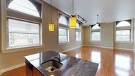 This single-floor space showcases a thoughtfully designed open-concept layout that seamlessly connects the kitchen and living areas. The kitchen is anchored by a striking dark granite island countertop with an undermount stainless steel sink, offering both functionality and visual appeal. Rich hardwood flooring flows throughout, adding warmth and continuity across the entire space.

Distinctive yellow pendant lights hang above the kitchen island, providing focused task lighting while contributing a bold design accent. A track lighting system complements the overhead illumination, ensuring the space is well-lit throughout. A ceiling fan is visible in the living area, enhancing comfort year-round.

Multiple windows dressed with horizontal blinds line the walls, inviting generous amounts of natural light and offering views of the surrounding greenery. The neutral, warm-toned walls serve as a versatile backdrop that complements both the cabinetry and flooring. Arched architectural window detailing adds a refined touch to the overall aesthetic.

The open floor plan creates a spacious, airy atmosphere that is ideal for both everyday living and entertaining. The combination of durable materials, modern fixtures, and classic architectural elements gives this space a timeless yet contemporary character.