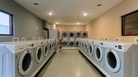 This dedicated laundry facility is designed to accommodate high-volume use with an impressive lineup of modern front-loading washing machines and dryers arranged in parallel rows along both sides of the room. The layout maximizes efficiency, providing easy access to each machine and ample space to move between units. Along the center of the room, stacked dryer units offer additional drying capacity, making the facility well-suited for serving a large number of users. The room features a polished terrazzo-style floor that is both durable and easy to maintain, while recessed lighting in the ceiling provides bright, even illumination throughout the space. The walls are finished in warm neutral tones, contributing to a clean and welcoming atmosphere. Each washer station includes individual control panels, and payment interfaces are conveniently positioned at each machine. Ventilation is well-addressed with ceiling vents and dedicated exhaust infrastructure visible in the space. The overall design is practical and functional, prioritizing accessibility and throughput while maintaining a tidy, organized environment suitable for everyday use.