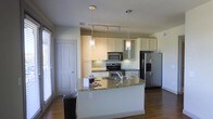 This single-floor space showcases a well-appointed kitchen as its central feature. The kitchen is designed with crisp white cabinetry that extends to the upper walls, offering ample storage through both open glass-front and solid-door cabinets. The countertops are finished in granite with warm, multi-toned flecks, providing both durability and visual interest. A generously sized kitchen island anchors the space, offering additional prep area and casual seating potential.

Stainless steel appliances, including a French door refrigerator with bottom freezer, a smooth-top range, and an over-the-range microwave, lend a sleek, contemporary feel. Two elegant glass pendant lights hang above the island, complemented by a track lighting system that ensures the workspace is well-lit throughout.

The kitchen opens to an adjacent living area, creating a connected, open-concept flow. Rich hardwood flooring runs throughout, adding warmth and continuity. Natural light filters in through a window fitted with blinds, brightening the overall atmosphere. The clean lines, neutral palette, and quality finishes combine to create a functional yet stylish living environment.