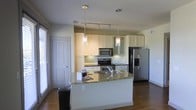This single-floor space showcases a well-appointed kitchen as its central feature. The kitchen is designed with crisp white cabinetry that extends to the upper walls, offering ample storage through both open glass-front and solid-door cabinets. The countertops are finished in granite with warm, multi-toned flecks, providing both durability and visual interest. A generously sized kitchen island anchors the space, offering additional prep area and casual seating potential.

Stainless steel appliances, including a French door refrigerator with bottom freezer, a smooth-top range, and an over-the-range microwave, lend a sleek, contemporary feel. Two elegant glass pendant lights hang above the island, complemented by a track lighting system that ensures the workspace is well-lit throughout.

The kitchen opens to an adjacent living area, creating a connected, open-concept flow. Rich hardwood flooring runs throughout, adding warmth and continuity. Natural light filters in through a window fitted with blinds, brightening the overall atmosphere. The clean lines, neutral palette, and quality finishes combine to create a functional yet stylish living environment.