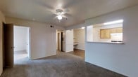 This single-floor residence offers a clean, functional layout designed for comfortable everyday living. The main living area is finished with wall-to-wall carpeting and neutral-toned walls, creating a warm and versatile backdrop. A ceiling fan with integrated lighting is centrally positioned, providing both air circulation and ambient light to the space. HVAC venting is conveniently located overhead, ensuring year-round climate control. A pass-through opening connects the living area to the kitchen, which features light wood cabinetry and a countertop bar — ideal for casual dining or entertaining. The kitchen's open sightlines into the living space enhance the overall sense of connectivity throughout the unit. Adjacent to the living area, a built-in desk nook with a dedicated countertop and shelving above offers a practical workspace or study area. The flooring transitions from carpet in the living space to hard surface in the adjoining areas, adding visual definition between zones. A hallway leads toward additional rooms, hinting at a well-organized floor plan beyond the main living space. Overall, the residence presents a tidy, move-in ready environment with practical built-in features and a cohesive, understated aesthetic.