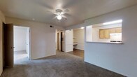 This single-floor residence offers a clean, functional layout designed for comfortable everyday living. The main living area is finished with wall-to-wall carpeting and neutral-toned walls, creating a warm and versatile backdrop. A ceiling fan with integrated lighting is centrally positioned, providing both air circulation and ambient light to the space. HVAC venting is conveniently located overhead, ensuring year-round climate control. A pass-through opening connects the living area to the kitchen, which features light wood cabinetry and a countertop bar — ideal for casual dining or entertaining. The kitchen's open sightlines into the living space enhance the overall sense of connectivity throughout the unit. Adjacent to the living area, a built-in desk nook with a dedicated countertop and shelving above offers a practical workspace or study area. The flooring transitions from carpet in the living space to hard surface in the adjoining areas, adding visual definition between zones. A hallway leads toward additional rooms, hinting at a well-organized floor plan beyond the main living space. Overall, the residence presents a tidy, move-in ready environment with practical built-in features and a cohesive, understated aesthetic.