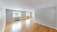 This single-floor residence is defined by its consistent use of warm, honey-toned hardwood flooring that flows seamlessly through the living, dining, and bedroom areas, creating a cohesive and inviting atmosphere. The layout, as revealed by the dollhouse view, is thoughtfully organized with distinct zones for living, sleeping, and cooking.

The expansive main living area is bright and open, benefiting from multiple windows fitted with white horizontal blinds that allow natural light to fill the space. Baseboard heating units run along the perimeter, and a ceiling fan with a built-in light fixture adds both comfort and practicality.

The bedroom is generously proportioned and features a ceiling fan, two closets, and a built-in vanity nook with a round mirror and granite countertop — a charming and functional detail. The neutral gray walls throughout the home lend a calm, modern backdrop.

The kitchen makes a bold statement with its classic black-and-white checkered tile floor, white cabinetry, granite countertops, white subway tile backsplash, and a stainless steel gas range with brass hardware accents. Two interior doors provide additional utility access.

The bathroom continues the timeless aesthetic with floor-to-ceiling white subway tile surrounding the bathtub, accented by a black border tile detail, a window with blinds, and hex tile flooring — a polished, classic finish.
