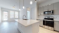 This contemporary single-floor unit is designed with a clean, modern aesthetic throughout, featuring light wood-style flooring and a crisp white-and-gray color palette that creates a bright, cohesive ambiance.

The open-concept kitchen serves as the heart of the space, anchored by a generously sized center island with a white quartz countertop, an undermount sink, and elegant pendant lighting hanging overhead. Gray flat-front cabinetry lines the walls, complemented by a marble-style tile backsplash and a full suite of stainless steel appliances, including a refrigerator with ice maker, electric range, built-in microwave, and dishwasher. The kitchen flows seamlessly into the living area, which is bathed in natural light from large windows and features a glass door opening to a private balcony with exterior views.

The bedroom is a calm, well-lit retreat with wide windows and neutral finishes. It is served by spacious walk-in closets equipped with multi-level shelving and hanging rods, offering generous storage capacity. The full bathroom features a floating gray vanity with a wide mirror, a soaking tub, and light tile flooring, all enhanced by a bright vanity light bar. The overall design prioritizes clean lines, natural light, and functional living throughout.