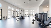 Located at 72 Eagle Rock Ave in East Hanover, NJ, this fitness center offers a generously sized, single-floor exercise space designed to accommodate a comprehensive range of workout needs. The room features soaring ceilings and a clean, modern aesthetic defined by crisp white walls, recessed lighting, and a durable dark epoxy-style flooring that spans the entire area.

Large windows positioned along multiple walls — including a striking row of clerestory windows near the ceiling — flood the space with natural light, creating an open and energizing atmosphere. The layout is thoughtfully organized to separate different training zones. The cardio section is anchored by a row of treadmills and elliptical machines, each equipped with individual screens, positioned directly in front of the large windows. A dedicated strength training area houses a multi-station cable machine, a flat bench, and a selection of selectorized resistance machines. A free weights zone features a dumbbell rack and additional functional training accessories, including a BOSU ball.

Full-length mirrors line one wall, visually expanding the space while allowing users to monitor their form. Built-in shelving provides convenient storage, and glass-paneled entry doors add a polished, contemporary touch. The overall design is bright, clean, and functional, making it a well-rounded facility for fitness enthusiasts.