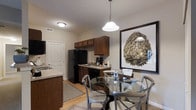 Located at 1524 Braxton Pky in Columbus, Ohio, this single-floor residence offers a well-designed living space with a cohesive and functional layout. The kitchen is outfitted with rich, dark wood cabinetry, black appliances including a full-sized refrigerator and range, and light-toned countertops that provide a clean contrast to the warm cabinetry. A kitchen island adds additional prep and counter space, enhancing the room's practicality. The light hardwood flooring runs throughout the kitchen and dining areas, contributing to a bright and welcoming atmosphere. Adjacent to the kitchen, the dining area is anchored by a round glass-top table surrounded by contemporary metal chairs, creating an intimate yet stylish setting. A large-scale botanical wall art piece serves as a bold focal point, adding a decorative touch to the space. A pendant light with a classic dome shade hangs above the dining area, providing warm, focused lighting. The overall ambiance of the space is casual yet refined, with neutral wall tones and thoughtful décor choices that create a comfortable and inviting environment throughout.