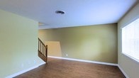 This two-story space offers a straightforward and functional layout with a clean, neutral aesthetic. The main living area is finished with warm hardwood-style flooring that extends throughout, providing a cohesive and grounded feel underfoot. The walls are painted in a muted olive-gold tone, offering a warm, earthy backdrop that pairs well with the white baseboards and trim detailing along the perimeter of the room.

A wooden staircase positioned toward one side of the main floor provides access to the upper level. The staircase features a classic wood railing with vertical balusters, adding a traditional touch to the space. The textured popcorn ceiling overhead includes a central HVAC vent, indicating a ducted climate control system is in place throughout the home.

The open feel of the main floor allows for flexible furniture arrangements, making it adaptable for various living configurations. The combination of warm wall tones, wood-style flooring, and clean white trim gives the space a modest yet inviting character. Overall, the two-floor layout provides practical separation between living areas, offering both openness on the main level and additional space above.