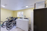 Located at 1550 Detroit St in Denver's Congress Park neighborhood, this shared utility room serves as both a laundry facility and storage area. The space is finished with soft yellow walls and light tile flooring, creating a clean, functional environment. At its core, the laundry area houses commercial-grade top-loading washer and dryer units positioned along one wall, accompanied by a water heater with exposed copper piping — a practical, utilitarian feature that speaks to the room's working character. A multi-shelf storage unit provides organized space for laundry detergents and cleaning supplies, keeping essentials readily accessible. A separate section of the room functions as dedicated bike storage, with a wooden rack accommodating multiple bicycles, including standard and electric models. A barred window along one wall allows natural light to filter into this area. Overhead, a flush-mounted ceiling light ensures even illumination throughout. A keyed entry door with signage provides secure access to the space. The overall layout is straightforward and efficient, designed to meet the practical day-to-day needs of building residents with clearly designated zones for laundry and bicycle storage.