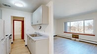 This well-organized single-floor unit offers a practical and comfortable layout across its thoughtfully arranged rooms. The galley-style kitchen runs along one wall, featuring light grey cabinetry — both upper and lower — a stainless steel sink with white countertops, and full-size appliances including a refrigerator and stove. Light hardwood-style flooring runs consistently throughout the unit, lending a clean and cohesive appearance.

The open living area is bright and airy, anchored by a large picture window with a warm wood sill that invites natural light and offers views of the surrounding neighborhood. A baseboard heating system runs beneath the windows, providing efficient warmth throughout. The clean white walls and textured ceiling create a neutral, versatile backdrop.

The bedroom is a comfortable and functional space, also featuring the same light flooring, a baseboard heater, a window with an outdoor view, and a warm wood-toned door. The room offers ample wall space for furnishing flexibility.

The bathroom is clean and straightforward, featuring a full-size soaking tub with an overhead shower, a wall-mounted sink, tile flooring, and a wood-framed mirror, all finished in crisp white tones that contribute to a bright and tidy atmosphere throughout.